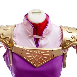 Puede incluir: Un disfraz de cosplay con una parte superior rosa y morada con detalles dorados. El disfraz incluye una armadura de hombro dorada adornada y una pieza de pecho dorada con un dise&ntilde;o geom&eacute;trico. El disfraz se exhibe en un maniqu&iacute;.