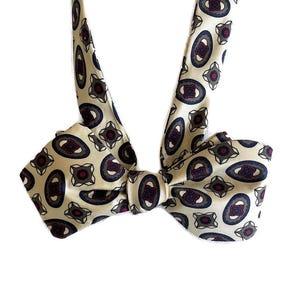 May include: A cream-colored bow tie with a repeating pattern of navy blue and burgundy ovals and geometric shapes. The bow tie is knotted and ready to wear, perfect for formal events or adding a touch of elegance to an outfit.