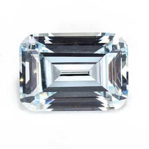 May include: A rectangular, light blue gemstone with an emerald cut. The gem has a clear, transparent appearance with multiple facets that reflect light. The gemstone is set against a white background.