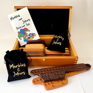 Può includere: Set di giochi di biglie e jolly in legno. Il set include una scatola di legno, tabelloni di gioco, sacchetti di velluto nero con scritte dorate e un libretto intitolato "Marbles and Jokers Rules and Tips". I tabelloni di gioco sono in legno chiaro e scuro.