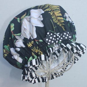 May include: A black shower cap with a floral magnolia design and a black and white checkered trim. The cap features a black and white polka dot bow and is displayed on a wire frame.