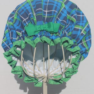 May include: A blue, green, and white plaid shower cap with a green bow and green polka dot trim. The cap is displayed on a white metal stand, showcasing its design and shape.