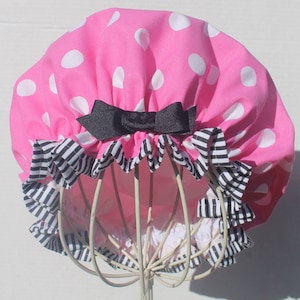 May include: A pink shower cap with white polka dots and a black bow. The cap has a ruffled edge with a black and white striped pattern. The cap is displayed on a white wire form.