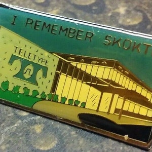 May include: A green and yellow enamel pin with the text "I Remember Skokte" and a depiction of a building with the text "Teletype" and the number "20" in a circle.