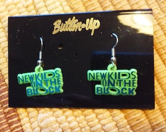 Könnte beinhalten: Ohrringe mit dem Text "NEW KIDS ON THE BLOCK" in Blau und Grün. Die Ohrringe befinden sich auf einer schwarzen Karte mit dem goldenen Text "Button-Up". Die Ohrringe haben silberne Haken.