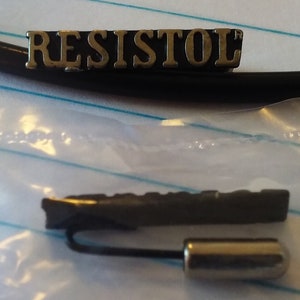 May include: A close-up of a black and silver Resistol brand hat pin. The pin is in a clear plastic bag. The pin has a black metal base with a silver metal top. The pin is about 2 inches long.