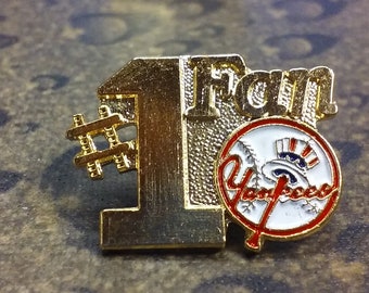 May include: Gold-colored pin with the number "1" and the word "Fan" above a white baseball with the New York Yankees logo. The number "1" has a hashtag symbol in front of it.