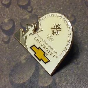 May include: A white enamel pin with a gold border. The pin features a stylized image of a runner, a mountain, and the Salt Lake 2002 Olympic Torch Relay logo. The text "Presenting Sponsor Chevrolet" is printed below the logo.