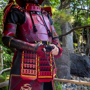 May include: A person wearing a red and black leather samurai armor with a skull design on the front. The armor is detailed with gold accents and black straps.