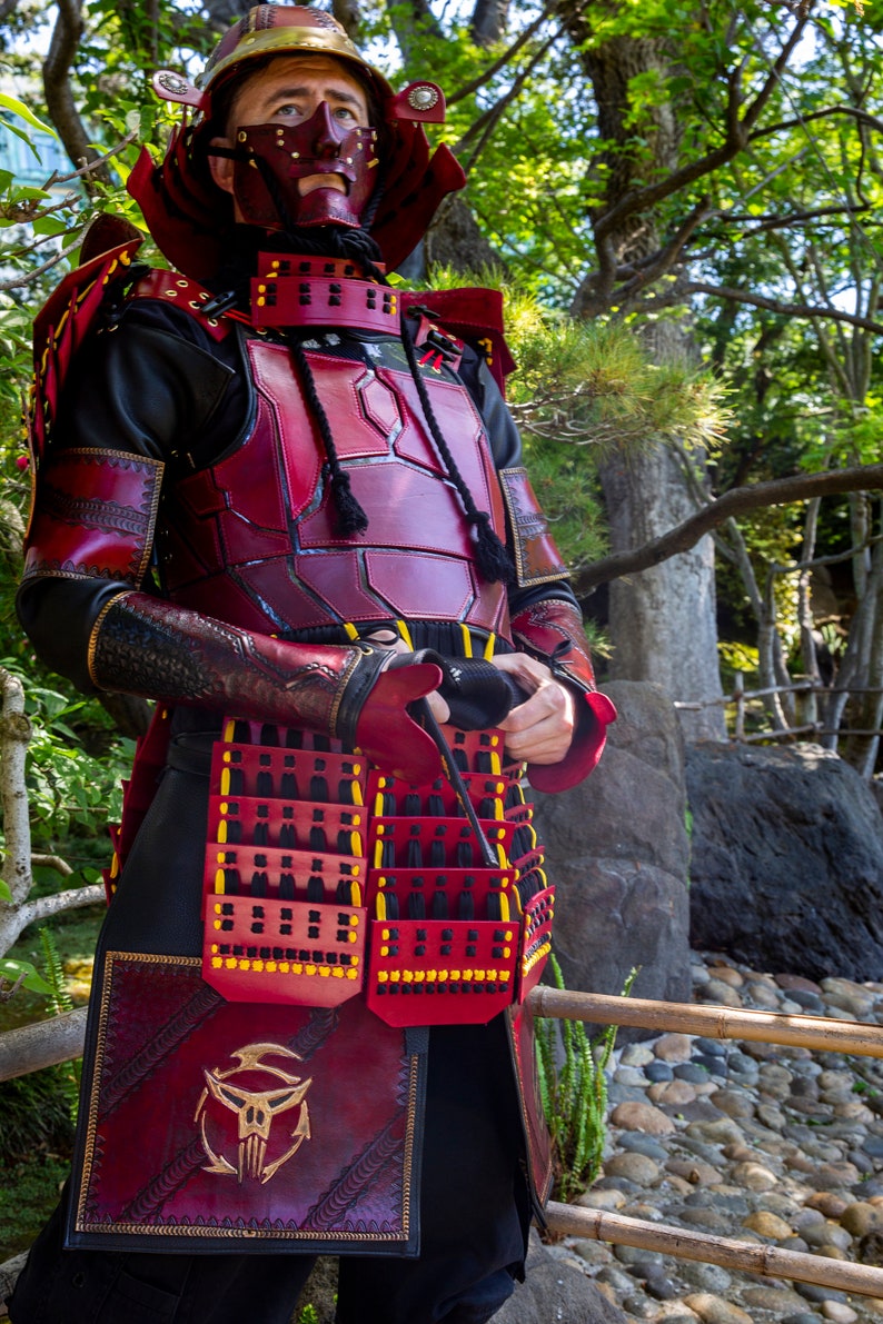 May include: A person wearing a red and black leather samurai armor with a skull design on the front. The armor is detailed with gold accents and black straps.