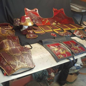 May include: A set of red and gold samurai armor, including a helmet, breastplate, and leg guards. The armor is decorated with intricate designs and is displayed on a table.