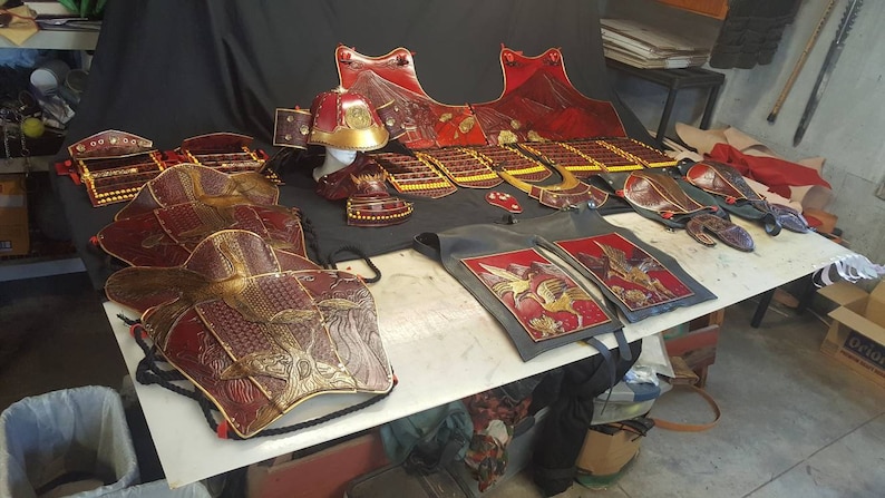 May include: A set of red and gold samurai armor, including a helmet, breastplate, and leg guards. The armor is decorated with intricate designs and is displayed on a table.