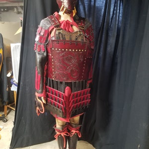 May include: A red and black leather samurai armor set with a dragon design. The armor includes a chest piece, skirt, and leg guards. The set is worn by a mannequin with a red and black mask.