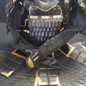 May include: Black leather samurai armor with gold accents. The armor includes a chest plate, shoulder pads, arm guards, and a skirt. The armor is displayed on a mannequin.