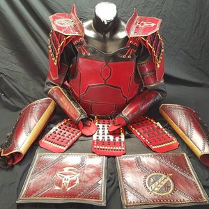 May include: A red and gold leather samurai armor set with intricate details. The armor includes a chest plate, shoulder pads, arm guards, and leg guards. The armor is displayed on a black background.