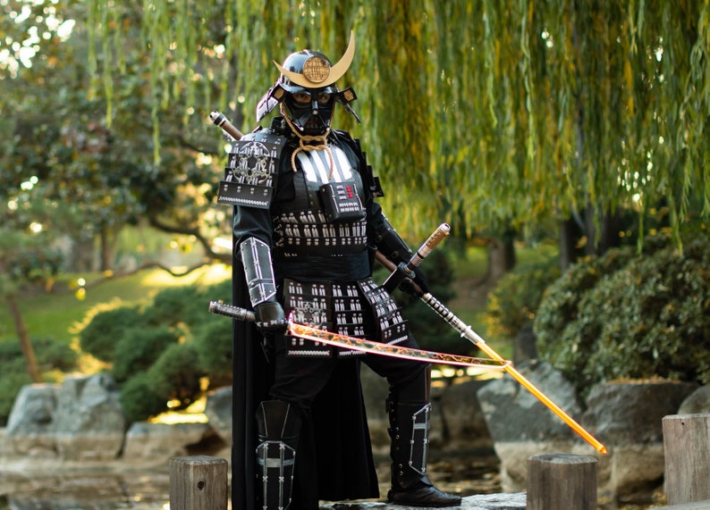 May include: A person dressed as Darth Vader in a black samurai costume with a gold helmet and a black cape, holding two lightsabers. The person is standing on a wooden platform in a garden setting.