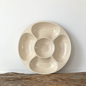 May include: A speckled beige ceramic serving platter with five compartments. The round platter has a central circular bowl surrounded by four oval-shaped sections. The surface has a speckled pattern, and the edges are slightly raised.