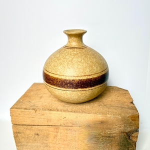 May include: A tan ceramic vase with a round body and a narrow neck. The vase features a wide, dark brown band around its center. It sits on a rustic, weathered wooden block, creating a contrast of textures and colors. The vase is a decorative item.