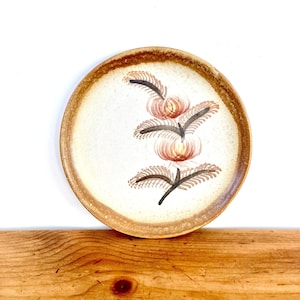 May include: A brown ceramic plate with a floral design. The plate has a white background with a brown rim and a stylized floral design in shades of brown, orange, and pink.