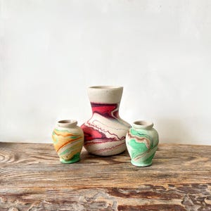 May include: A set of three ceramic vases with a marbled finish. The central vase is larger with a wide base and a neck, featuring swirls of deep red and white. Two smaller vases flank it, one with orange and green swirls and one with green and brown.