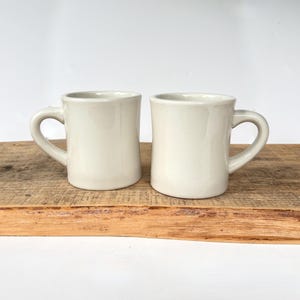 May include: Two matching off-white ceramic mugs with a smooth, glossy finish and a sturdy, rounded handle on each side. The mugs feature a classic cylindrical shape with a slightly tapered base, providing a clean and simple aesthetic for daily use.