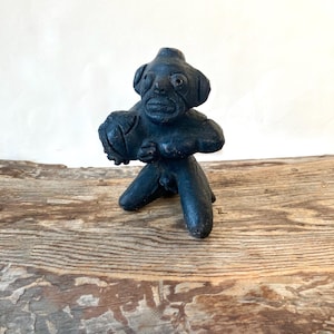 May include: A dark, weathered sculpture of a humanoid figure with a large head, holding a ball-like object. The figure is kneeling with its arms wrapped around the ball. The sculpture is on a wooden surface.