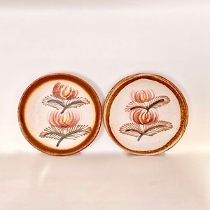 May include: Two small ceramic plates with a brown rim and a white center. Each plate features a hand-painted design of three stylized flowers in shades of pink, orange, and brown.