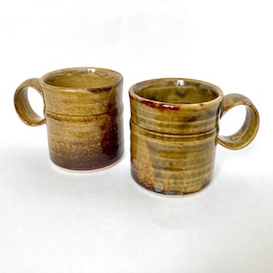 May include: Two brown and yellow ceramic mugs with a textured glaze. The mugs have a rounded shape and a handle.