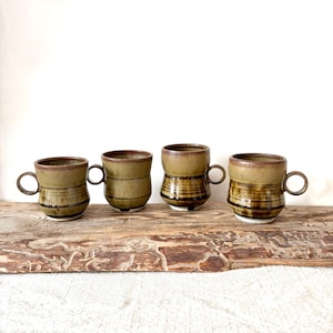 May include: Four handmade ceramic mugs with a unique design. The mugs are a mix of olive green and brown with a textured, striped pattern. Each mug has a small, rounded handle and a dark brown rim. They are displayed on a rustic wooden surface.