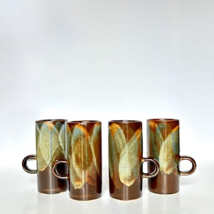 May include: Four tall, brown ceramic mugs with a unique glaze pattern of gold, green, and brown. Each mug has a small, rounded handle. The mugs are cylindrical and sit on a white surface.