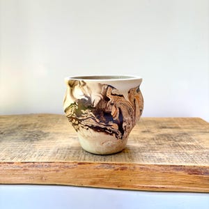 May include: A small, rounded ceramic planter featuring a cream-colored base with swirling, marbled patterns in shades of dark brown, black, and hints of orange. The vessel has a smooth, wide rim and a tapered base, showcasing a fluid, organic design.