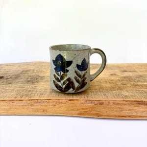 May include: A ceramic mug with a speckled light gray finish, featuring a hand-painted design of two dark blue flowers with green and brown stems and leaves. The mug has a cylindrical body, a sturdy handle on the right side, and a smooth rim.