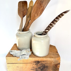 May include: Wooden kitchen utensils, including spatulas and spoons, stand in two off-white ceramic jars. A decorative feather is in one jar. The jars sit on a rustic wooden block, with a piece of bark on the block.
