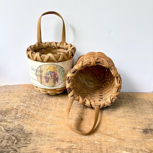 May include: Two round woven light brown baskets with a single arched handle each. One basket stands upright and features a paper label with the text Doorknob Basket and a floral illustration. The second basket is tilted on its side.