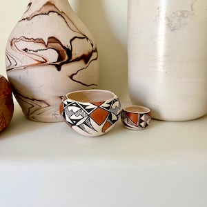 May include: A still life featuring two small, hand-painted ceramic bowls with geometric designs in black, white, and terracotta. The bowls are displayed alongside two decorative vases, one with a marbled pattern and the other a solid white.