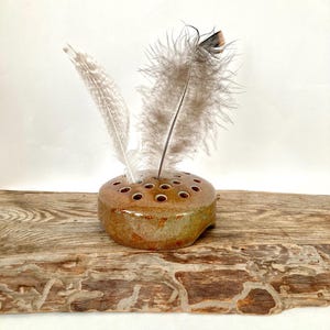 May include: A small, brown ceramic bud vase with multiple holes for stems. The vase is sitting on a piece of weathered wood. Two white feathers are in the vase.