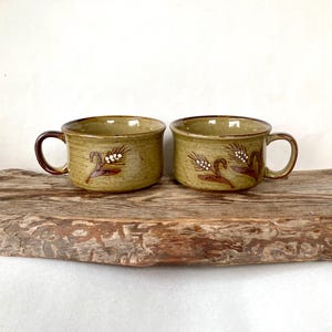 May include: Two ceramic mugs with handles. The mugs are a muted green color with a brown rim and handle. Each mug features a wheat stalk design in brown and white. The mugs are sitting on a wooden surface.