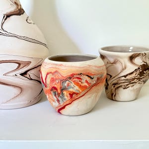 May include: A collection of three ceramic vessels featuring marbled patterns. One large vase and two smaller bowls display swirling designs in shades of cream, brown, orange, and red, with smooth, rounded forms and open tops.