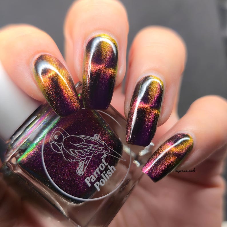 May include: A bottle of Parrot Polish nail polish with a purple and gold iridescent finish. The nails are painted with the same color-shifting polish, showcasing a cross-like design. The bottle has a white cap and a label with a parrot illustration.