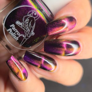 May include: Close-up of a hand with long, oval-shaped nails painted with a color-shifting nail polish. The polish transitions from purple to gold and orange. The bottle of Parrot Polish is visible in the background.