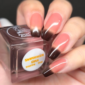 May include: A close-up of a bottle of Parrot Polish nail polish next to a hand with long, square-shaped nails. The nails are painted with a gradient effect, transitioning from a light pink to a dark brown color. The nail polish bottle has a brown cap and a white label.