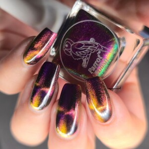 May include: A hand displays long, oval-shaped nails painted with a color-shifting nail polish. The polish transitions from gold to pink to purple. A square bottle of Parrot Polish is held in the hand, with a parrot logo on the label.