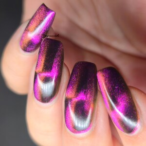 May include: Close-up of manicured nails with a cat-eye effect. The nails are painted with a shimmering polish that transitions from purple to orange and black, creating a unique and eye-catching design. The nails are oval-shaped.