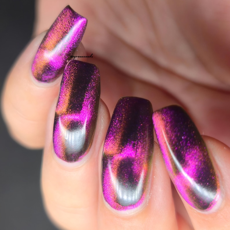May include: Close-up of manicured nails with a cat-eye effect. The nails are painted with a shimmering polish that transitions from purple to orange and black, creating a unique and eye-catching design. The nails are oval-shaped.