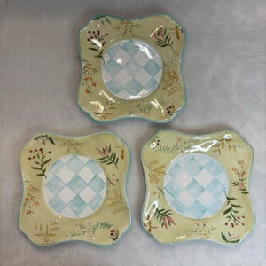 May include: Three square ceramic plates with scalloped edges, featuring a central light blue and white checkered pattern surrounded by a pale yellow border decorated with painted green leaves, brown stems, and small red berries.