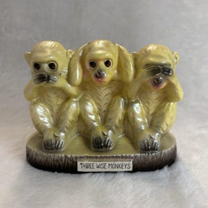 May include: A ceramic figurine of three monkeys sitting on a base with a wood-grain texture. The monkeys are light yellow with dark accents. One covers its mouth, one its ears, and one its eyes. A white plaque reads THREE WISE MONKEYS.