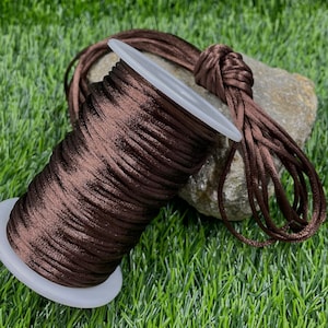 May include: A spool of dark brown satin cord sits on a white plastic base. The cord is tightly wound, with a portion unwound and knotted. The background is green, suggesting a natural setting. This cord is suitable for jewelry making and crafts.