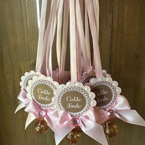May include: A collection of pink baby shower favors. Each favor features a scalloped-edged tag with the text "Cielito Lindo", a pink bow, a decorative gem, and a clear, round charm. The favors are suspended by pink ribbons.