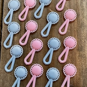 May include: A collection of plastic baby rattles in pastel pink and blue. Each rattle has a circular head with a scalloped edge and a loop handle. The rattles are arranged on a wooden surface.