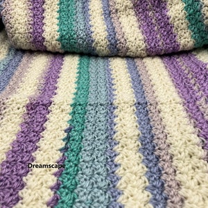 May include: A handmade crochet blanket with a striped pattern in shades of cream, purple, teal, and blue. The blanket is folded over, showcasing the texture and color variations. The word "Dreamscape" is visible on the blanket.